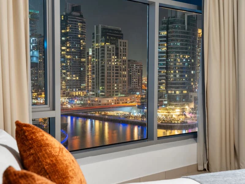 A made hotel bed with a rust cushion, a marina of lit towers filling the window beyond it at night