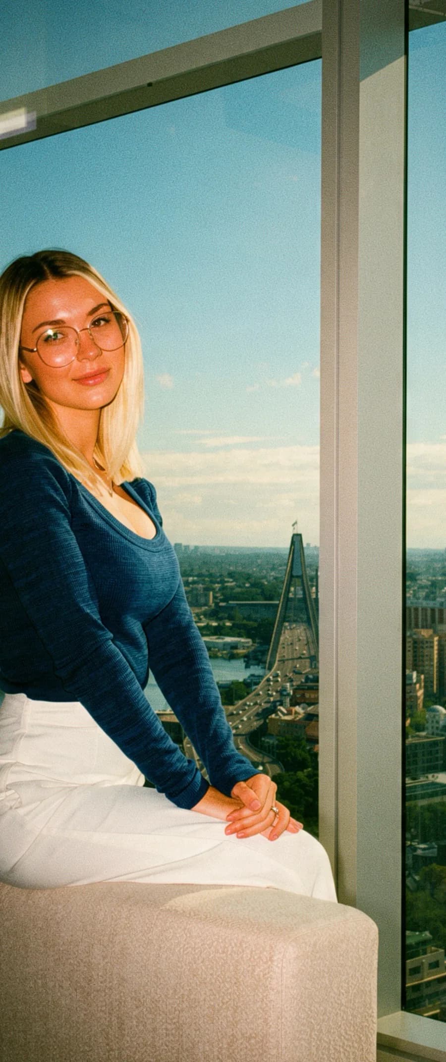 A woman seated by floor-to-ceiling glass high above the city, the bridge and the harbour below