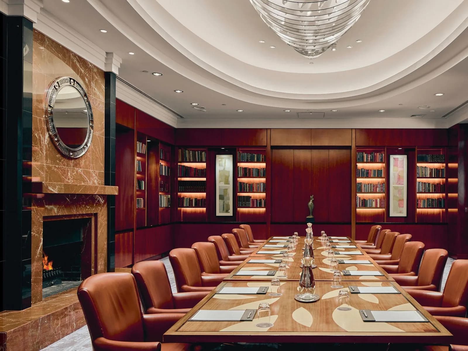 A private boardroom set for twenty: tan leather chairs down a long table, lit bookshelves, a fire and a round mirror on the left