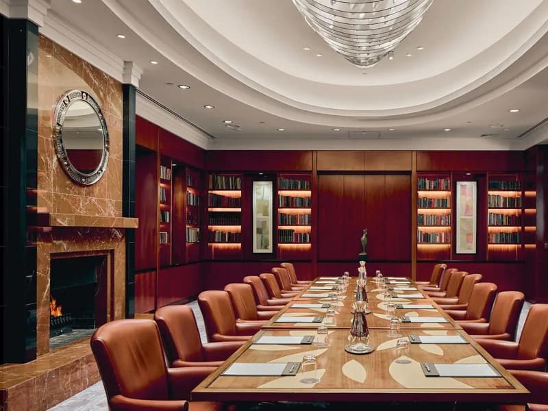 A private boardroom set for twenty: tan leather chairs down a long table, lit bookshelves, a fire and a round mirror on the left