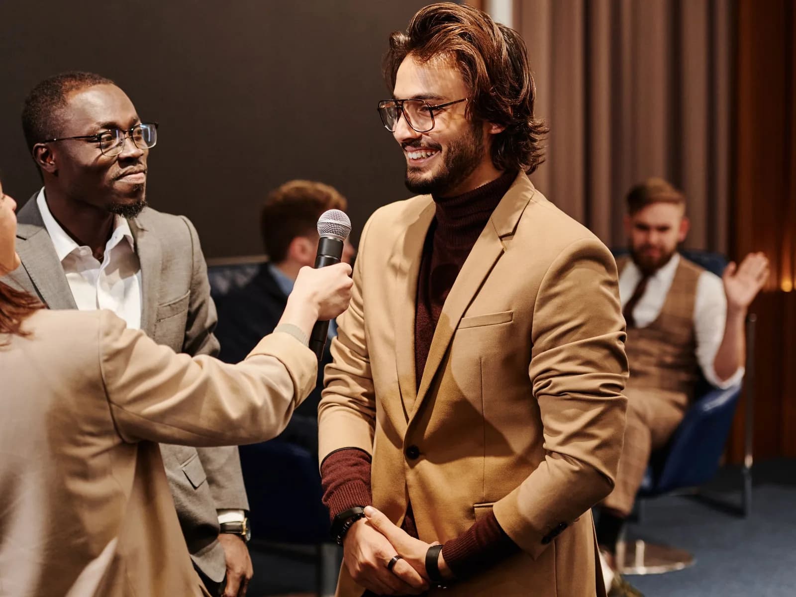 A guest in a tan jacket smiles as he is interviewed with a microphone at an event