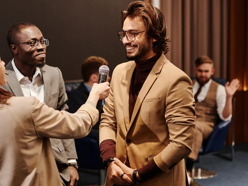 A guest in a tan jacket smiles as he is interviewed with a microphone at an event