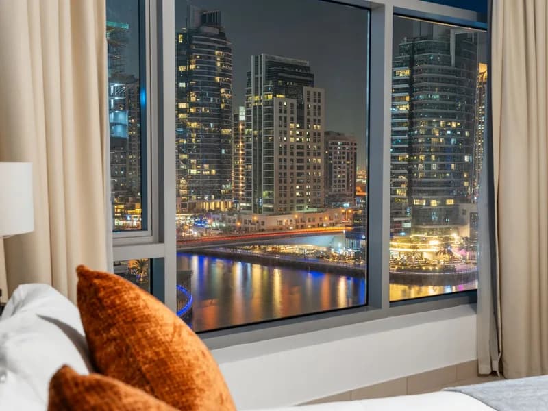 A made hotel bed with an orange cushion, a city of lit towers and a marina beyond the window at night