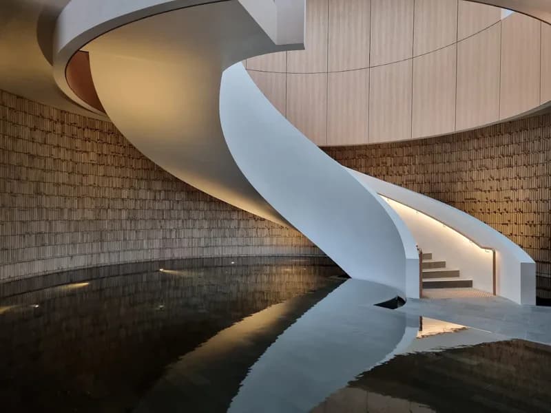 A white spiral staircase curving down over a still indoor pool, the water holding its reflection
