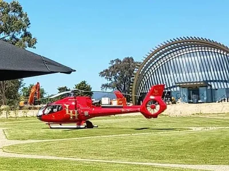 A red helicopter on the lawn at a winery, the cellar door and the vines behind it