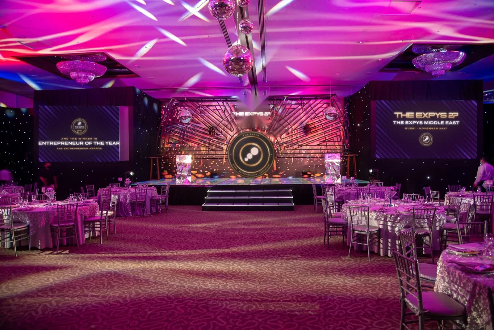 A violet-lit ballroom with a stage between two screens showing The Expys Middle East