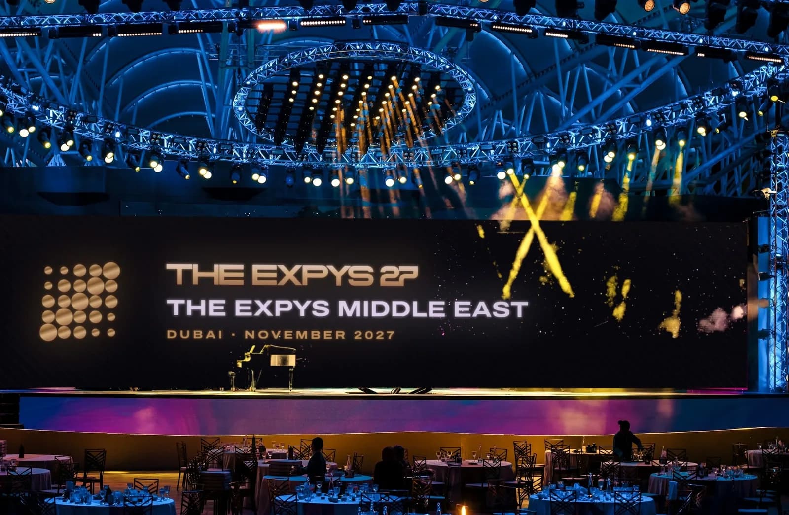 A stage under a lit truss dome, one LED wall the width of the stage showing THE EXPYS 27 and The Expys Middle East, rounds of ten below