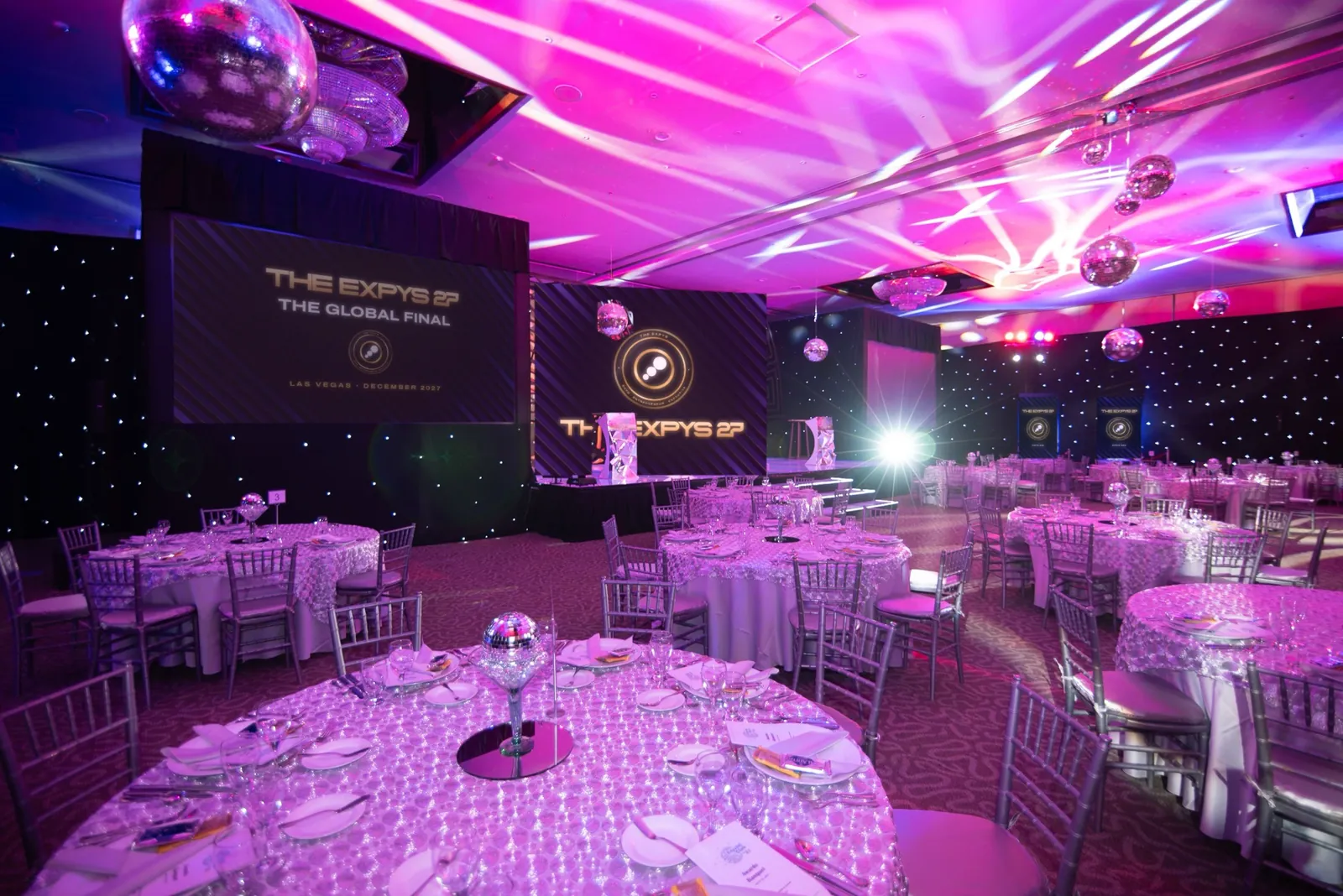 A violet-lit ballroom set for dinner, THE EXPYS 27 and The Global Final on the screen beside the stage