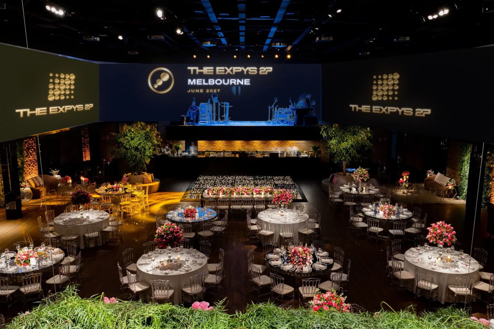 A room wrapped in screens over rounds of ten, THE EXPYS 27 and Melbourne above the stage