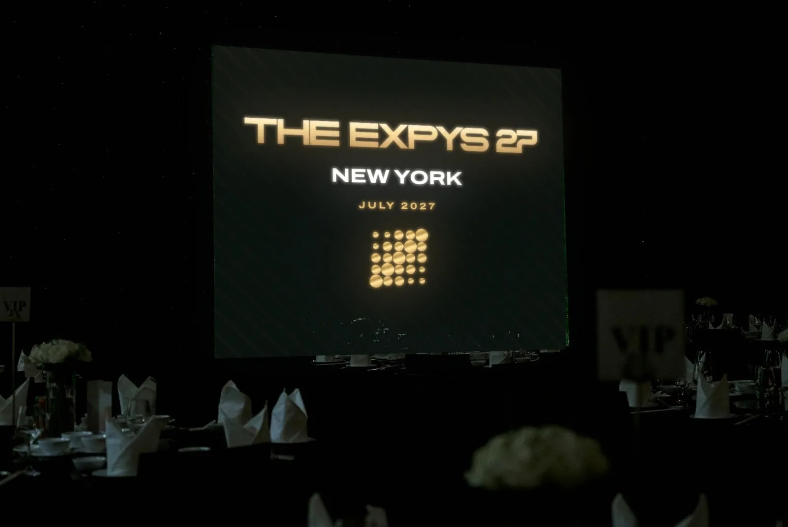 A dark room of dressed rounds, THE EXPYS 27 and New York on one big screen over the tables
