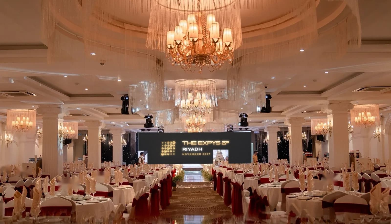 A white-columned ballroom under fringed chandeliers, THE EXPYS 27 and Riyadh on the LED wall over the stage