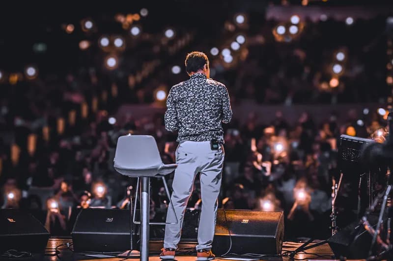 A speaker on stage seen from behind, the audience lit by their phones