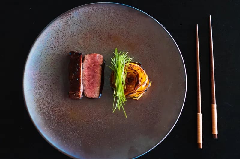 Seared beef, sliced to show its pink centre, with shaved greens and a roasted vegetable rose on a dark plate