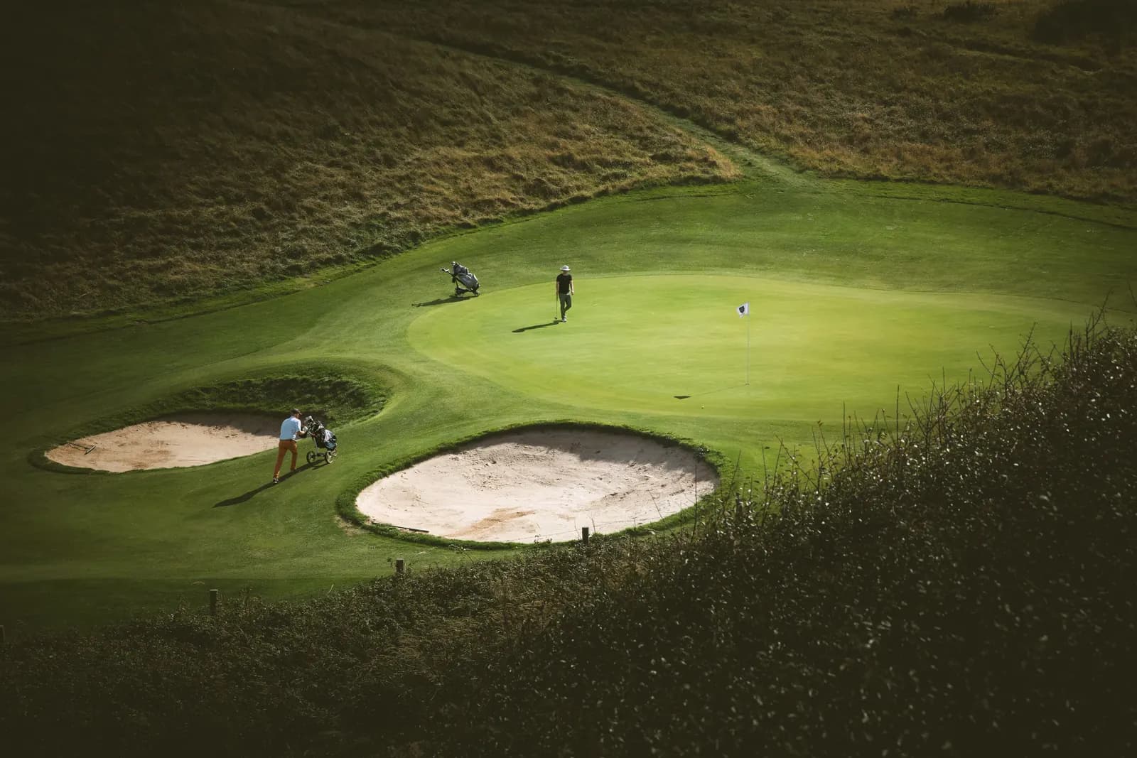 Golfers on a links green between two bunkers, seen from the hill above