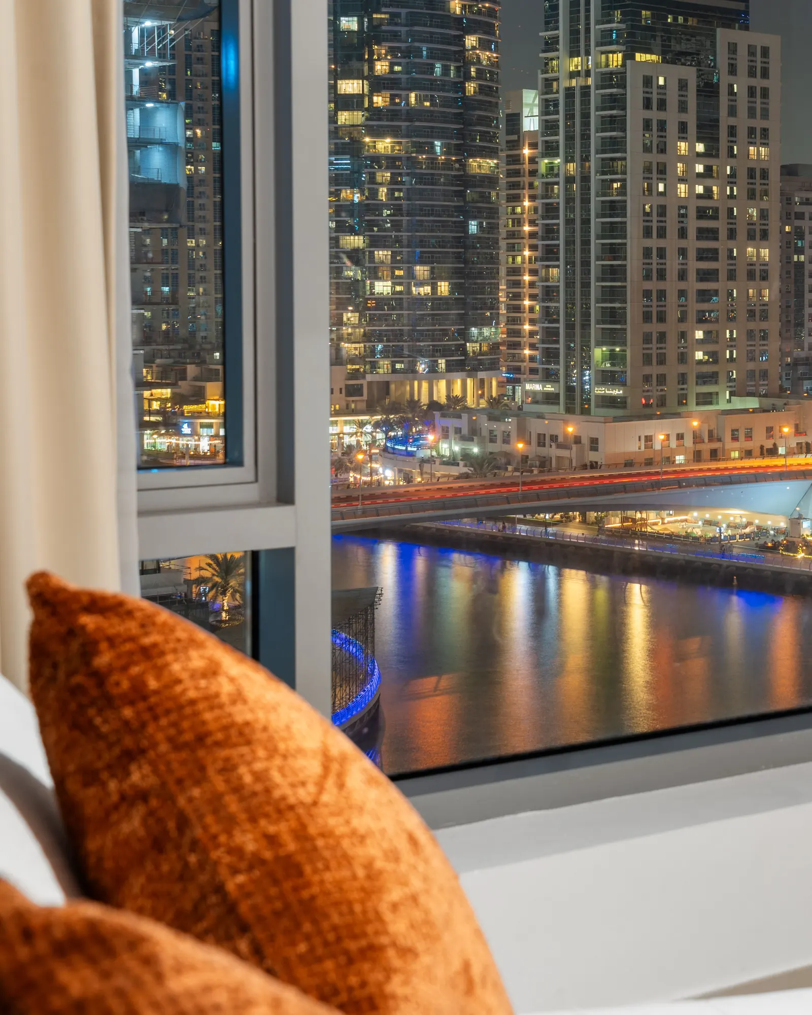 A hotel bed with a velvet cushion beside a tall window, lit towers and a waterway outside at night