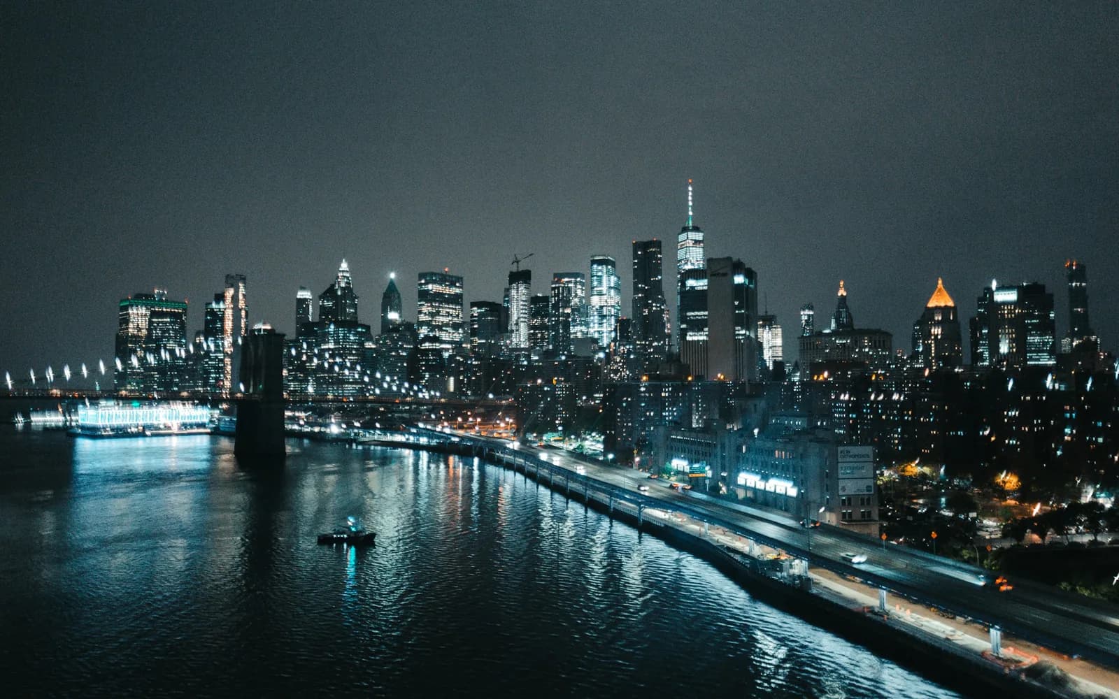 Lower Manhattan lit at night across the East River, the Brooklyn Bridge on the left